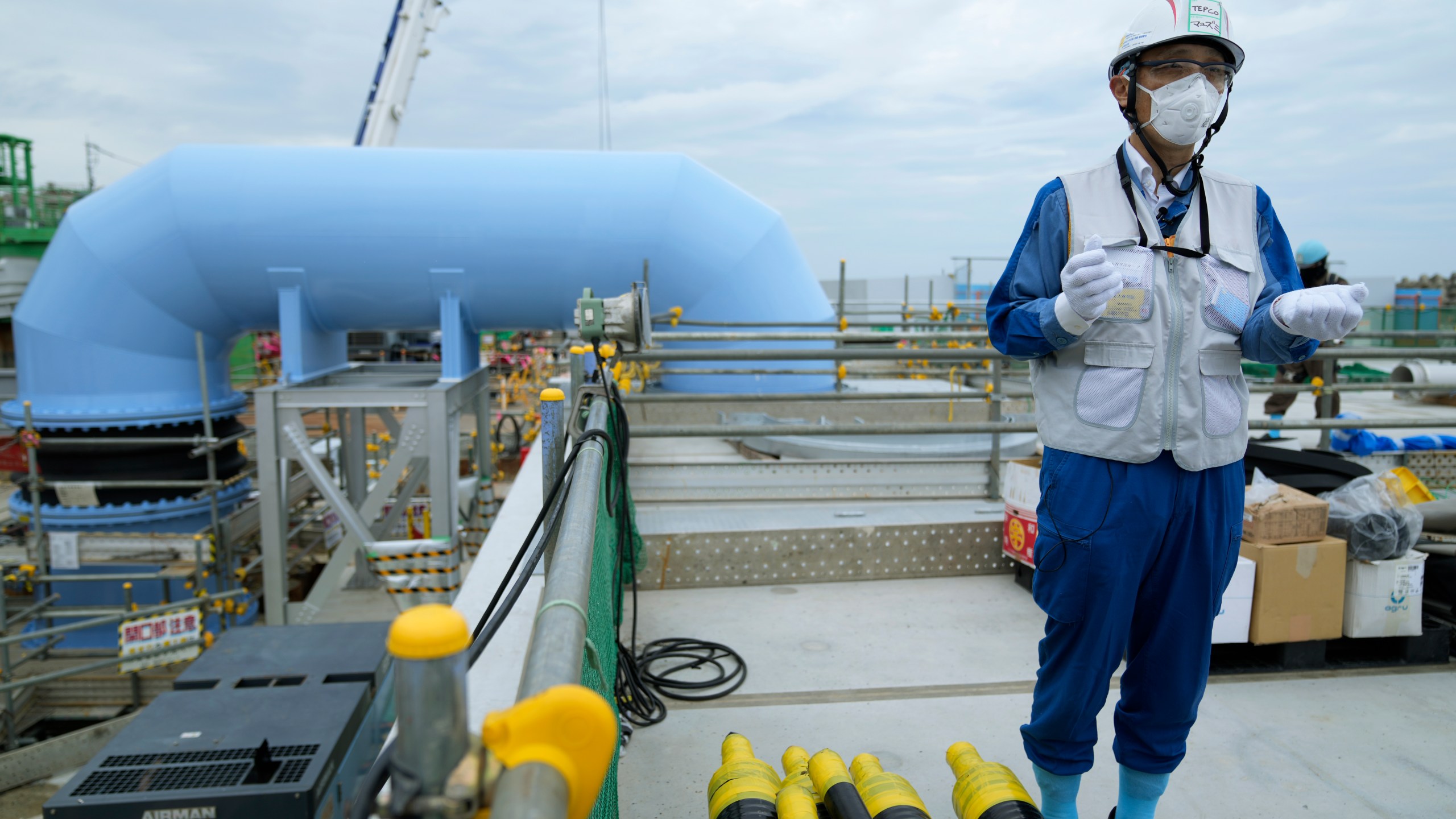 Tomohiko Mayuzumi, a spokesperson of Tokyo Electric Power Company Holdings, also known as TEPCO, speaks during a tour of the Fukushima Daiichi nuclear power plant for The Associated Press in Futaba town, northeastern Japan, Friday, July 14, 2023. He says treated radioactive water will be diluted with more than hundred times the seawater in this blue pipe, background, to levels much safer than international standards, before released into the sea. (AP Photo/Hiro Komae)