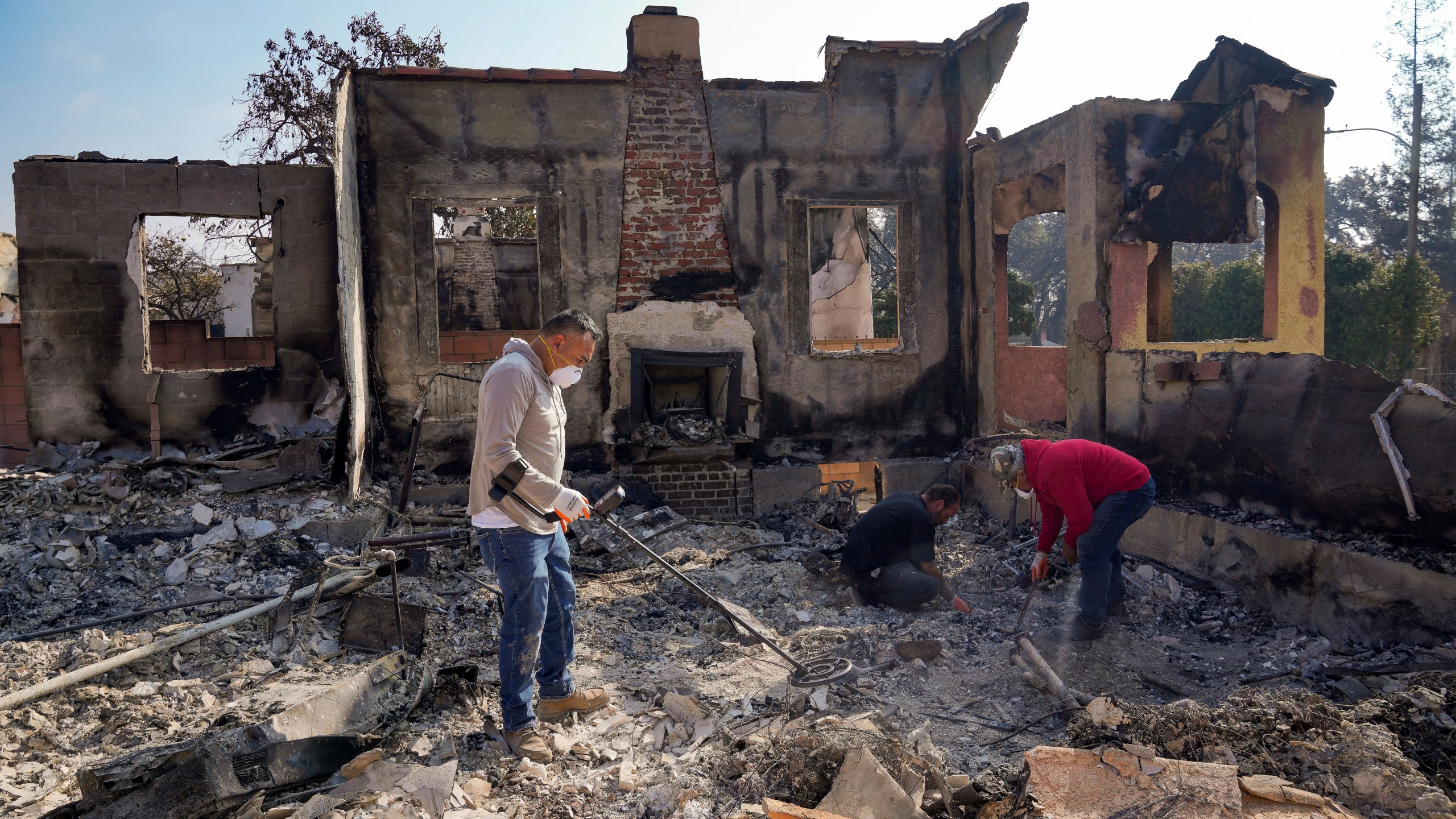 Homeowner David Marquez, left, holds a metal detector as his father, Juan Pablo Alvarado, right, and a friend look for the remains of gold jewelry and other silver items inside the walls of their multi-generational home in the aftermath of the Eaton Fire, Sunday, Jan. 19, 2025, in Altadena, Calif. (AP Photo/Damian Dovarganes)