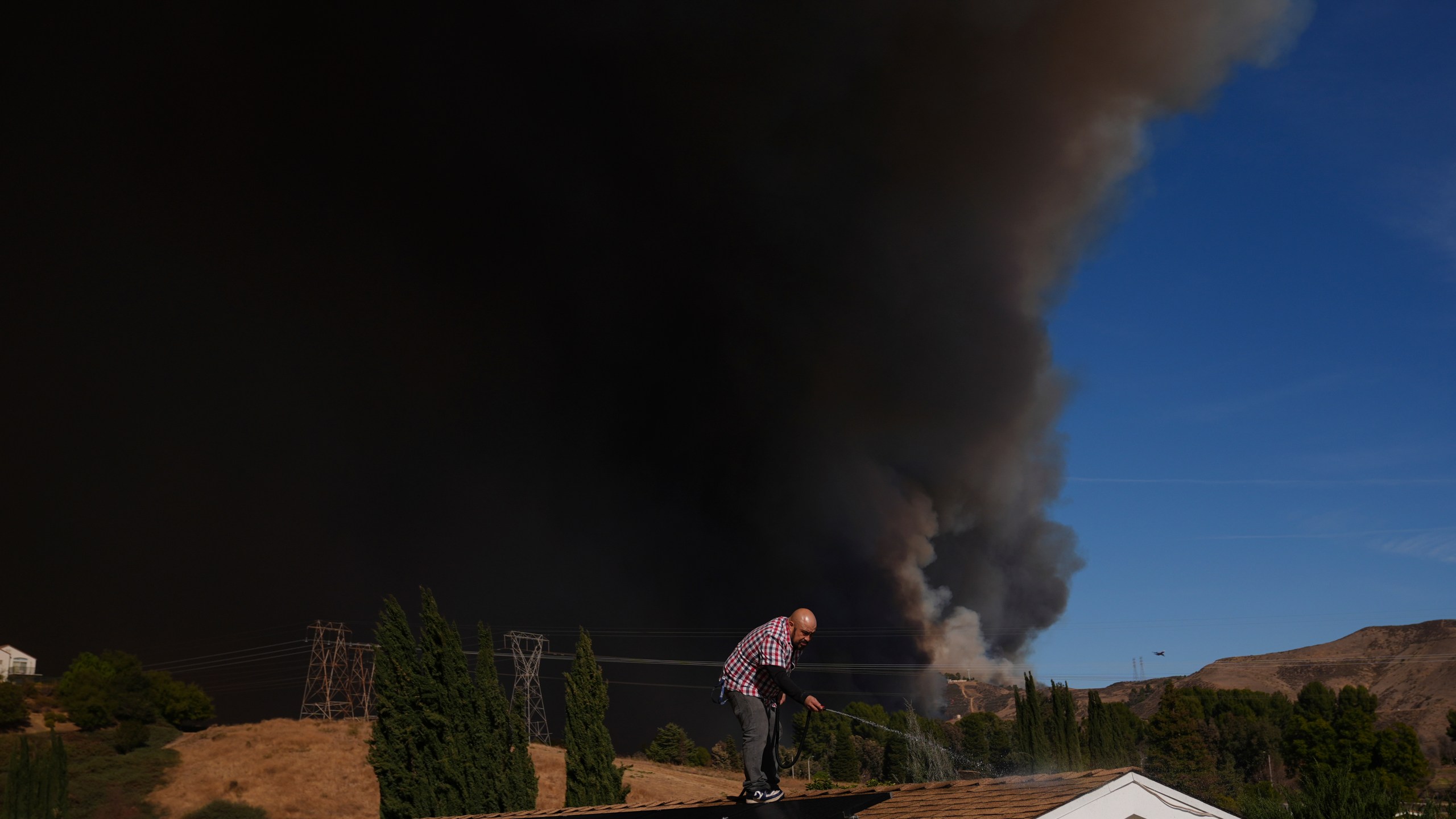 A home owner sprays water from the top of the roof in Castaic, Calif., as a large plume of smoke caused by the Hughes Fire rises from Castaic Lake Wednesday, Jan. 22, 2025. (AP Photo/Marcio Jose Sanchez)