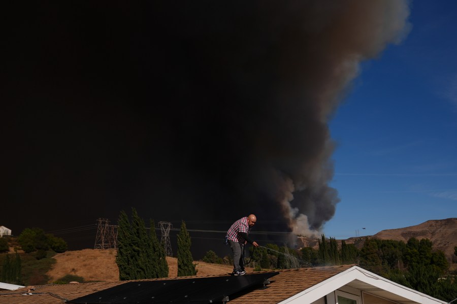 A home owner sprays water from the top of the roof in Castaic, Calif., as a large plume of smoke caused by the Hughes Fire rises from Castaic Lake Wednesday, Jan. 22, 2025. (AP Photo/Marcio Jose Sanchez)