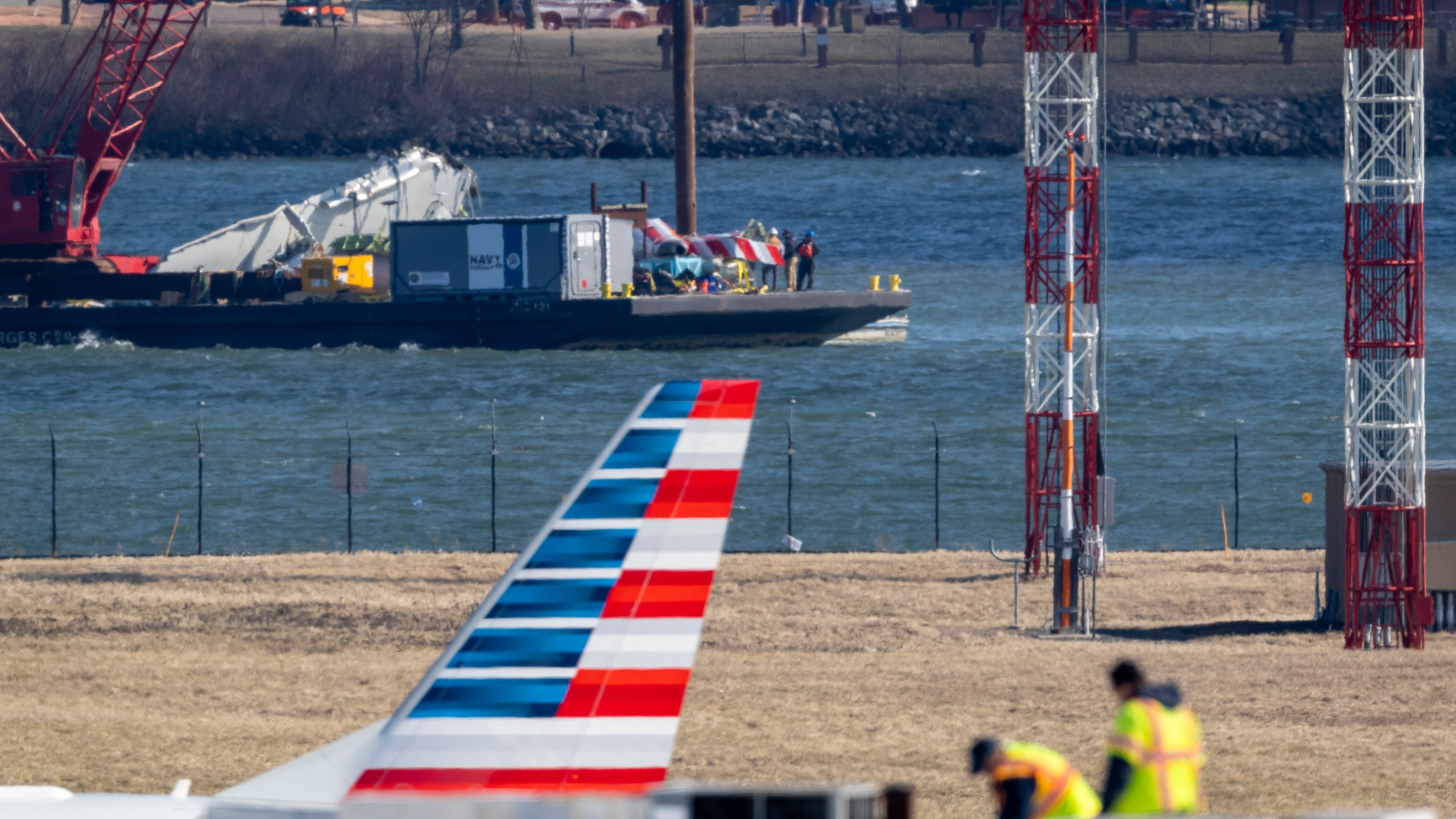 Parts of a plane are lifted from the water near the wreckage site in the Potomac River of a mid-air collision between an American Airlines jet and a Black Hawk helicopter, at Ronald Reagan Washington National Airport, Tuesday, Feb. 4, 2025, in Arlington, Va., as an American Airlines aircraft is seen landing on the runway, foreground. (AP Photo/Ben Curtis)