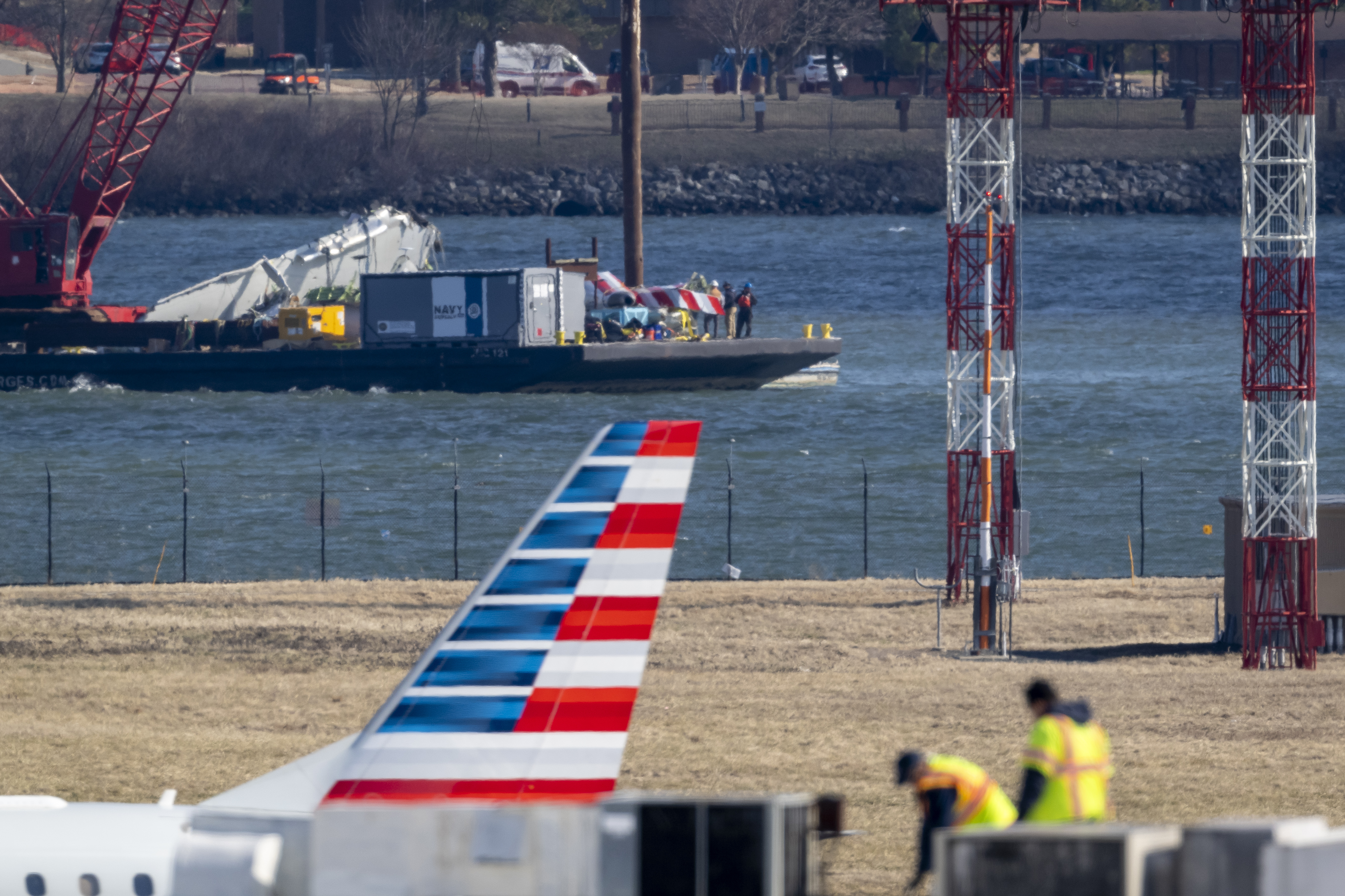 Parts of a plane are lifted from the water near the wreckage site in the Potomac River of a mid-air collision between an American Airlines jet and a Black Hawk helicopter, at Ronald Reagan Washington National Airport, Tuesday, Feb. 4, 2025, in Arlington, Va., as an American Airlines aircraft is seen landing on the runway, foreground. (AP Photo/Ben Curtis)