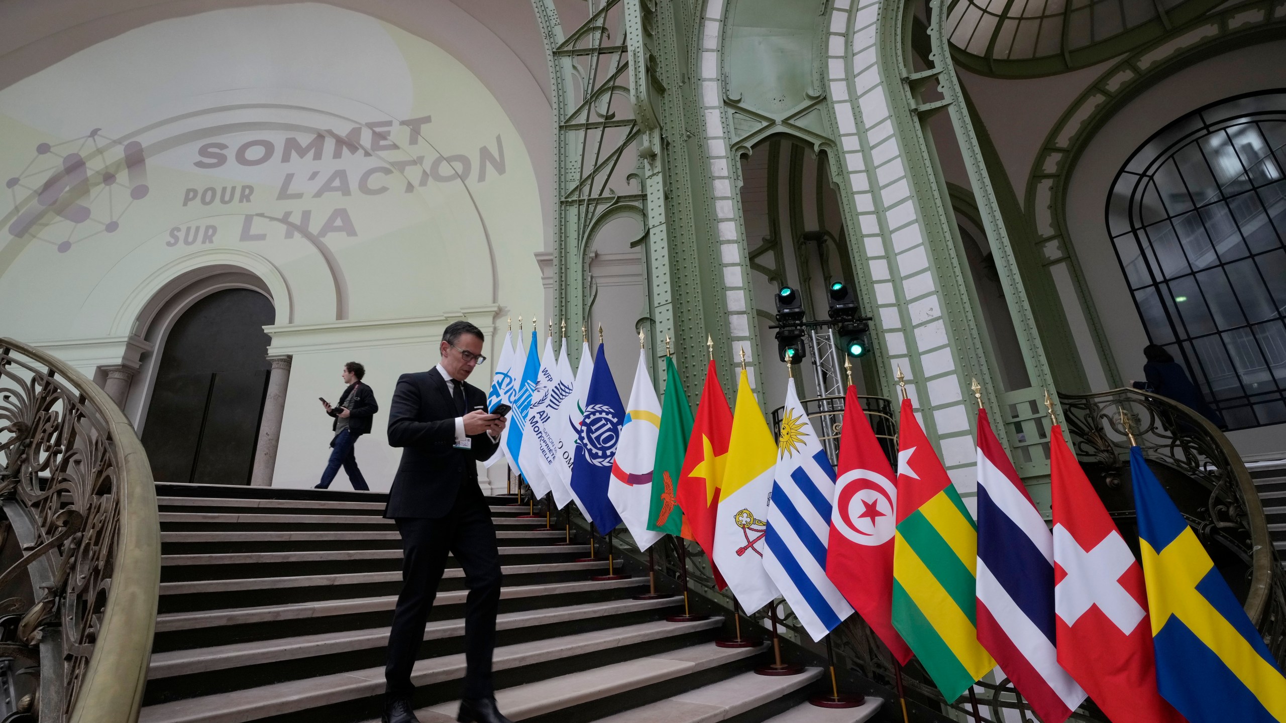 A man looks at his mobile phone as he walks past flags during an Artificial Intelligence Action Summit at the Grand Palais in Paris, Monday, Feb. 10, 2025. (AP Photo/Michel Euler)