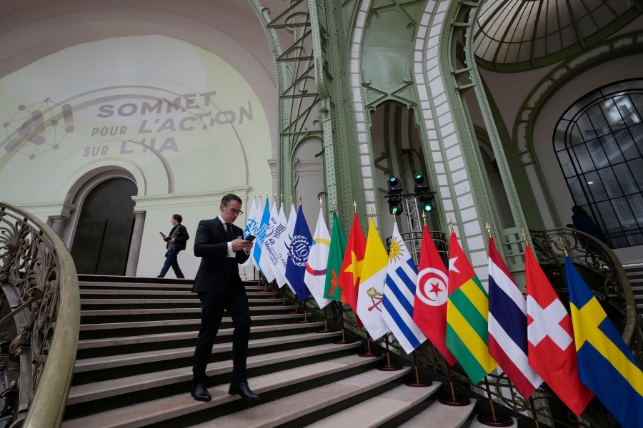 A man looks at his mobile phone as he walks past flags during an Artificial Intelligence Action Summit at the Grand Palais in Paris, Monday, Feb. 10, 2025. (AP Photo/Michel Euler)