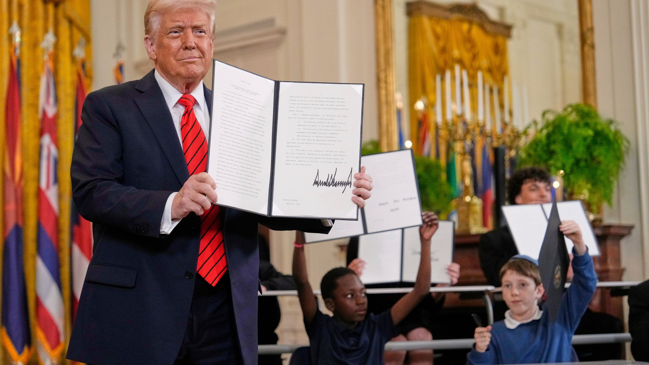 President Donald Trump, left, holds up a signed executive order as young people hold up copies of the executive order they signed at an education event in the East Room of the White House in Washington, Thursday, March 20, 2025. (AP Photo/Ben Curtis)